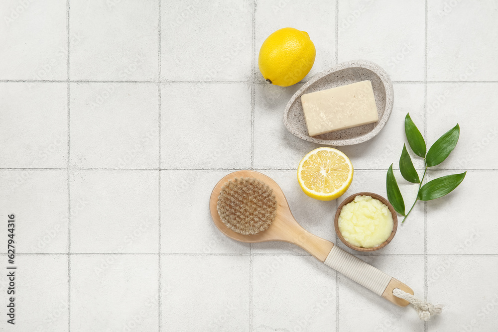 Bowl of lemon body scrub with massage brush and soap on white tile background