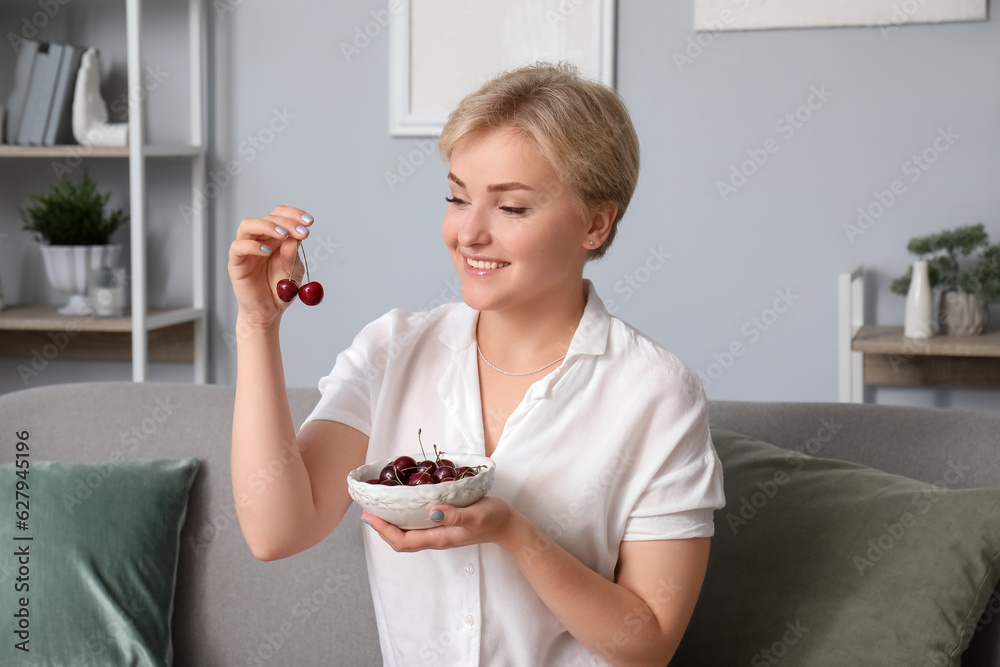 Beautiful happy young woman with bowl of ripe cherries sitting on sofa in living room