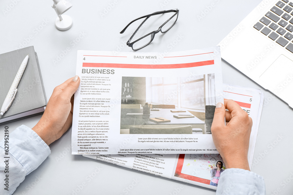 Woman reading newspaper at workplace with laptop and eyeglasses on grey background