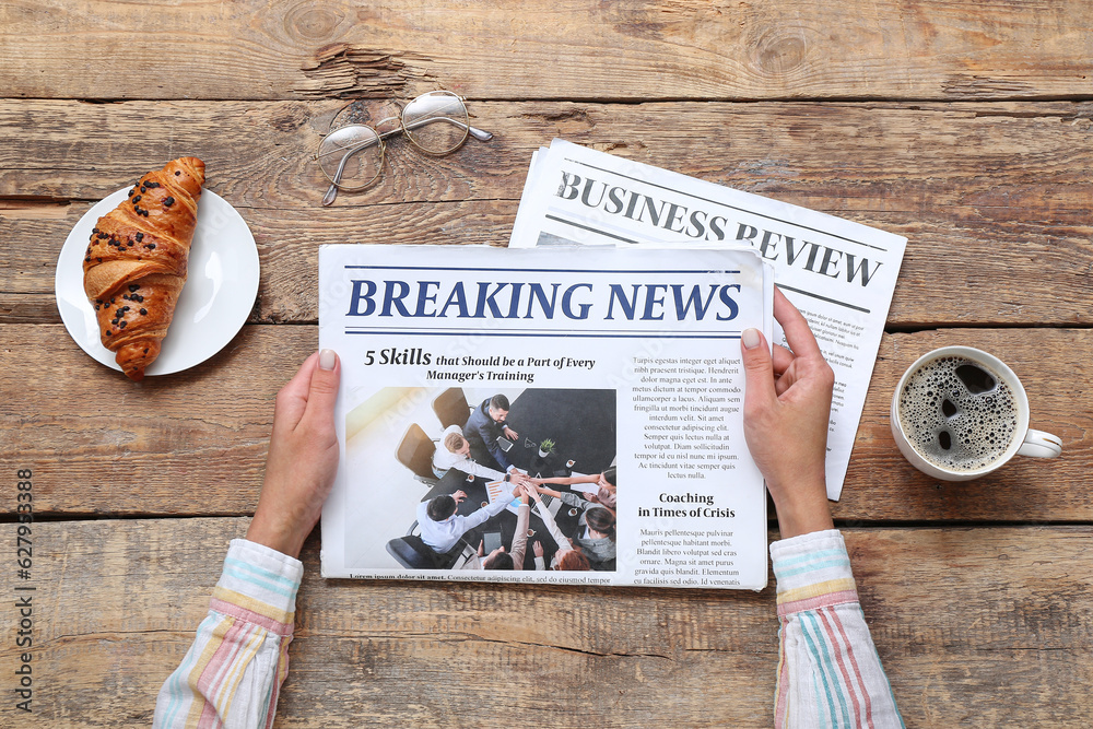 Woman reading newspaper with cup of coffee and croissant on wooden background