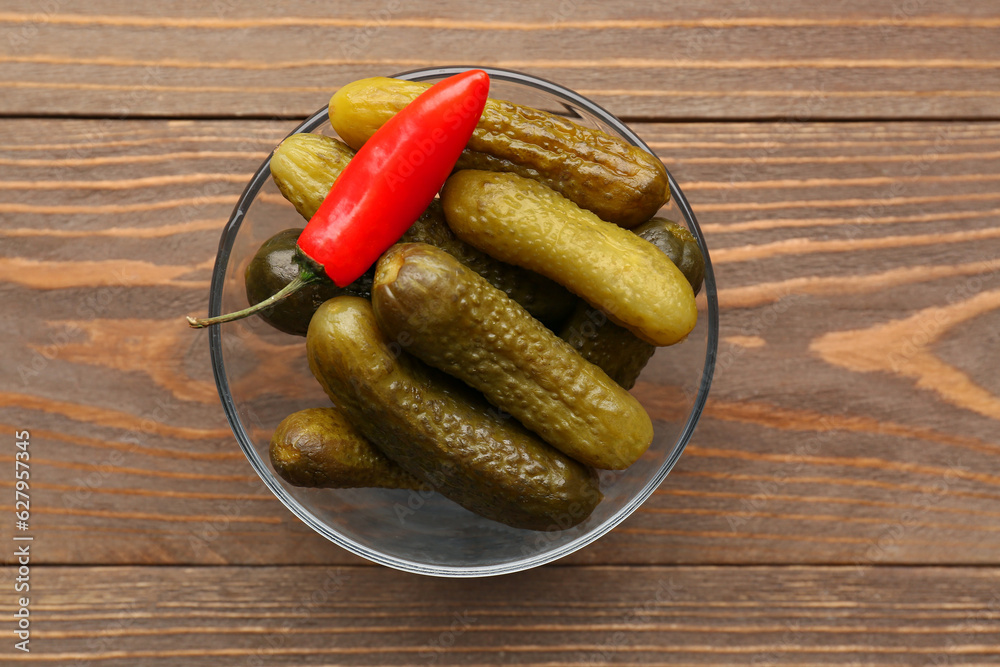 Glass bowl with pickled cucumbers and chili pepper on wooden background