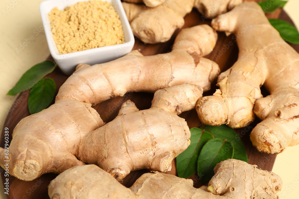 Wooden board with fresh ginger roots, leaves and bowl of dried powder on yellow background