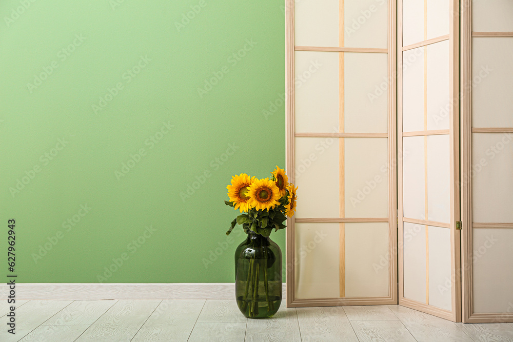 Vase with beautiful sunflowers and folding screen near green wall in room