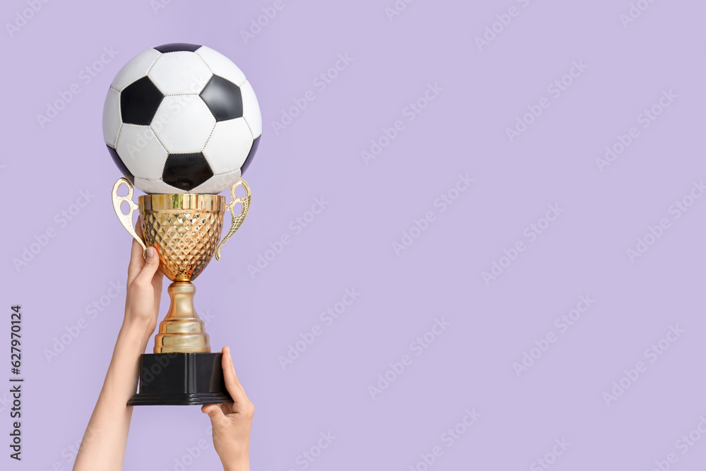 Female hands with gold cup and soccer ball on lilac background