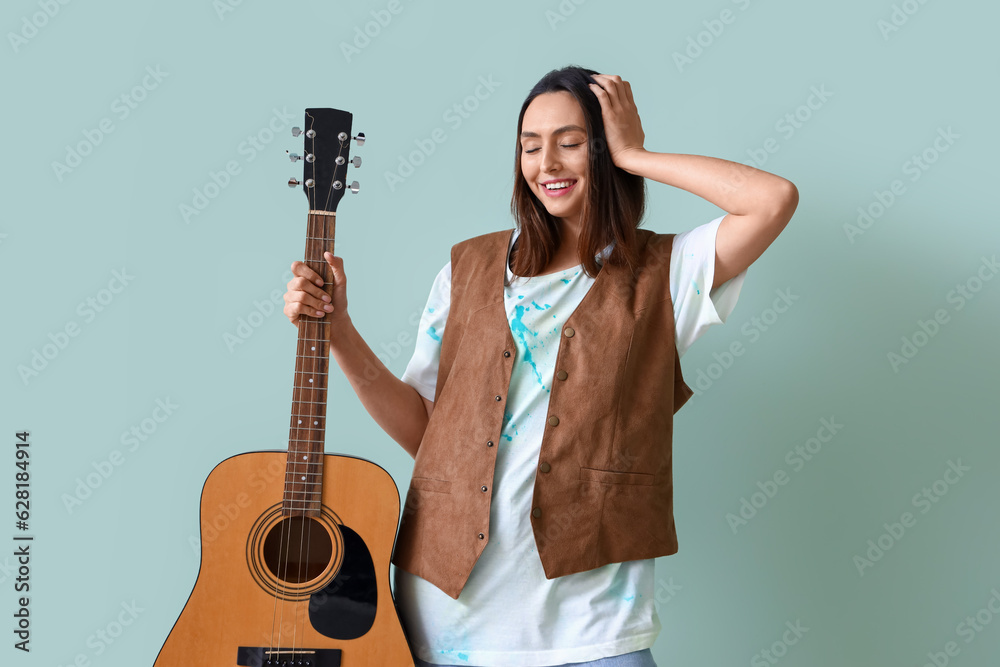 Stylish young woman in tie-dye t-shirt and with guitar on light blue background