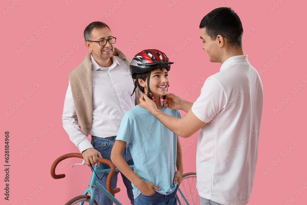 Young man putting bike helmet onto his little son and father on pink background