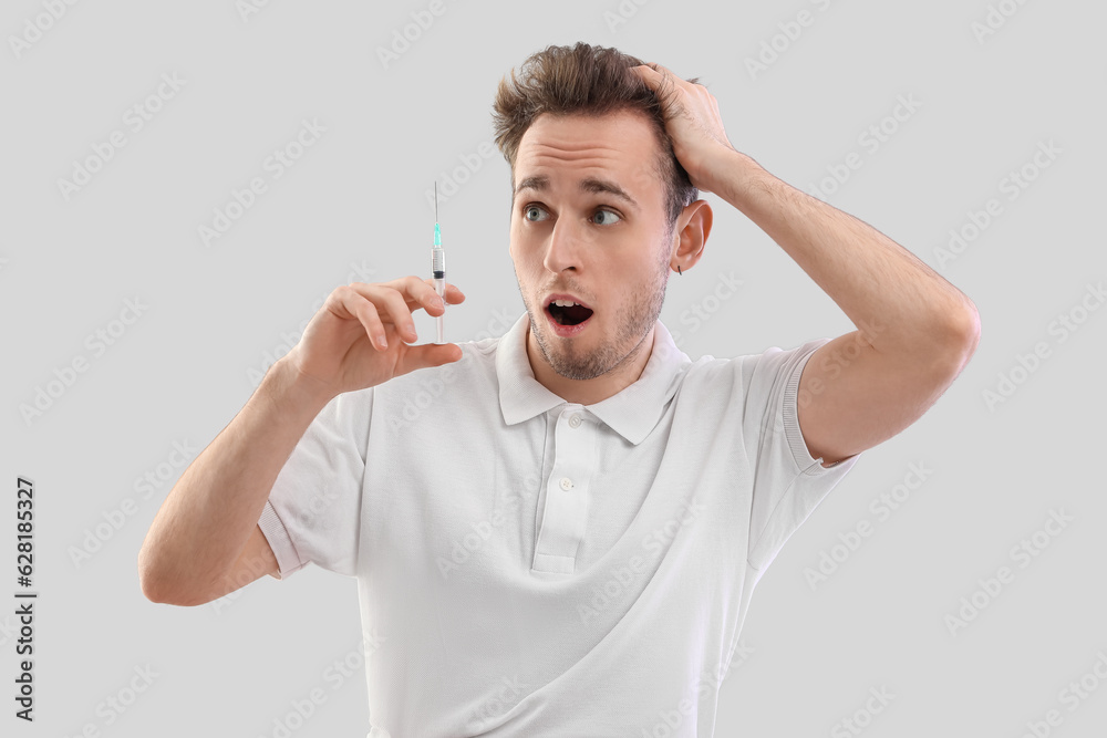 Shocked young man with injection for hair growth on grey background