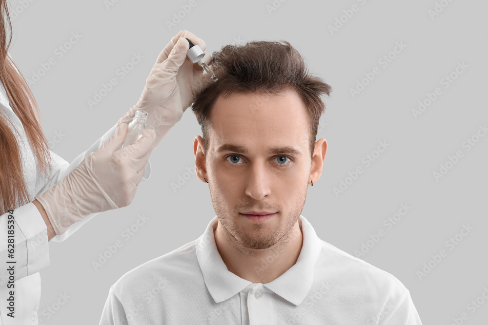 Young man and doctor with serum for hair growth on grey background