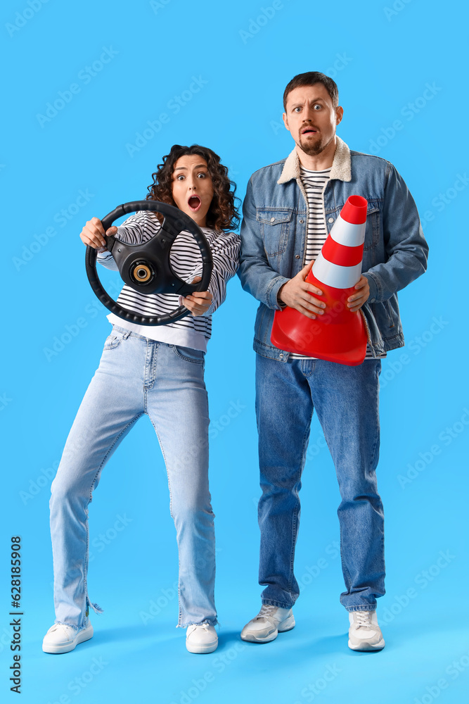 Shocked young couple with steering wheel and traffic cone on blue background