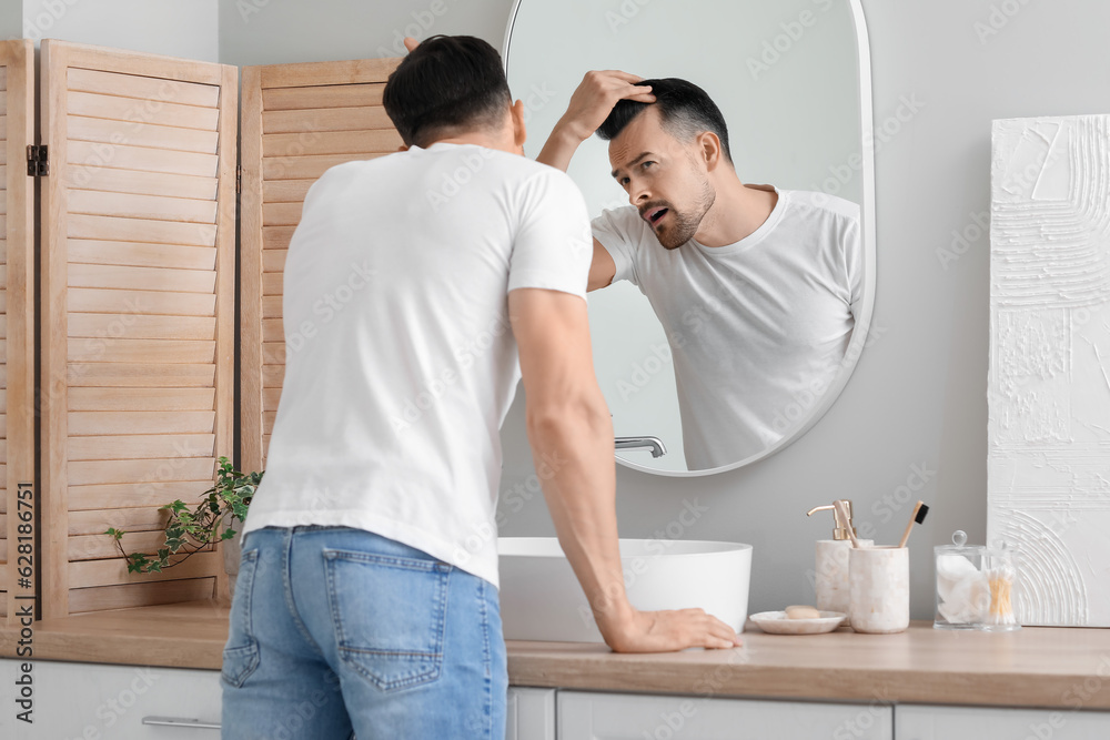 Worried young man with hair loss problem looking in mirror at home