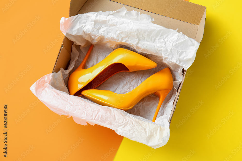 Cardboard box with high heeled shoes on color background