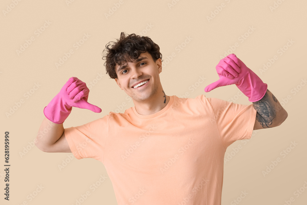 Young man in rubber gloves pointing at himself on beige background