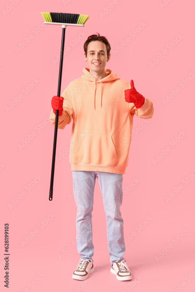 Young man with broom showing thumb-up on pink background