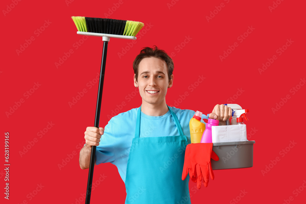 Male janitor with broom and bucket of cleaning supplies on red background