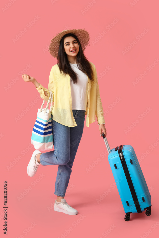 Young woman with beach bag and suitcase on pink background