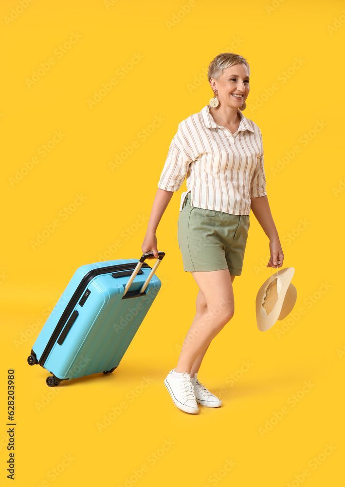 Mature woman with suitcase and summer hat on yellow background