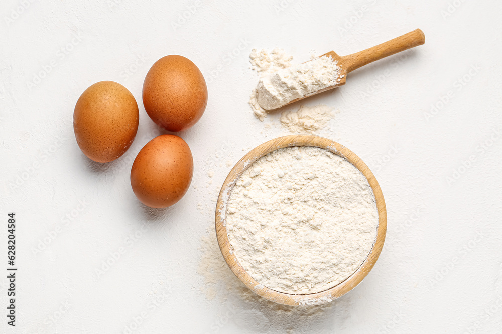 Wooden bowl with wheat flour, scoop and eggs on white background