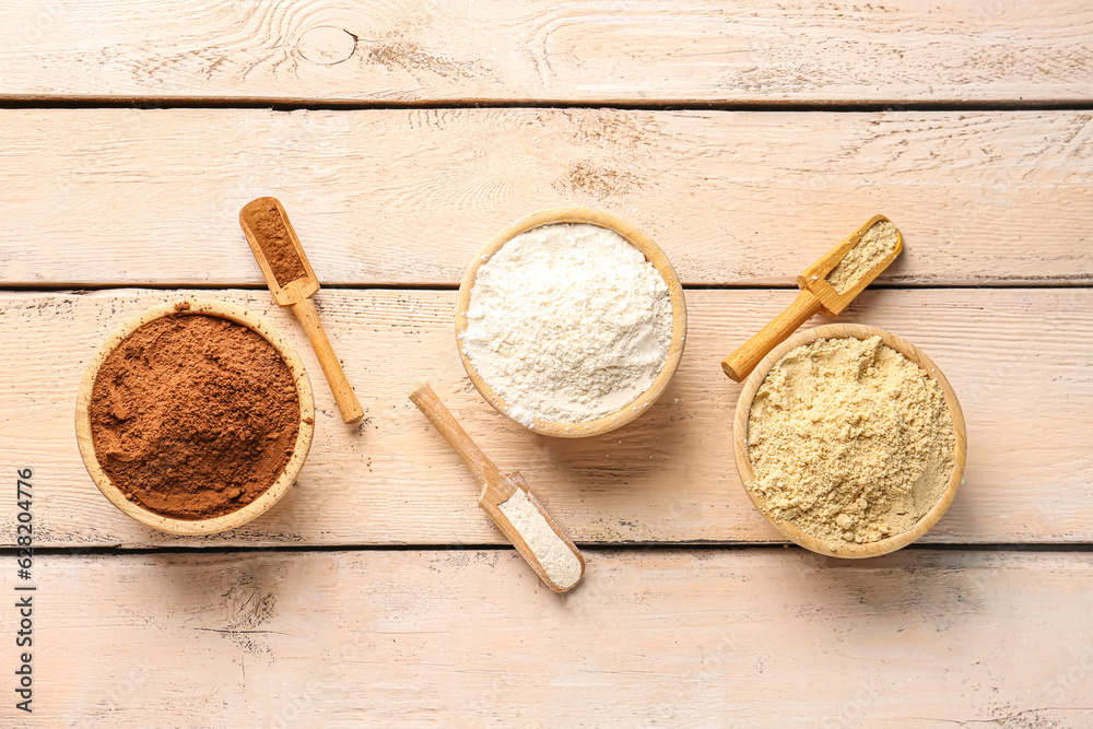 Wooden scoops and bowls with cacao, wheat and sesame flour on light wooden background