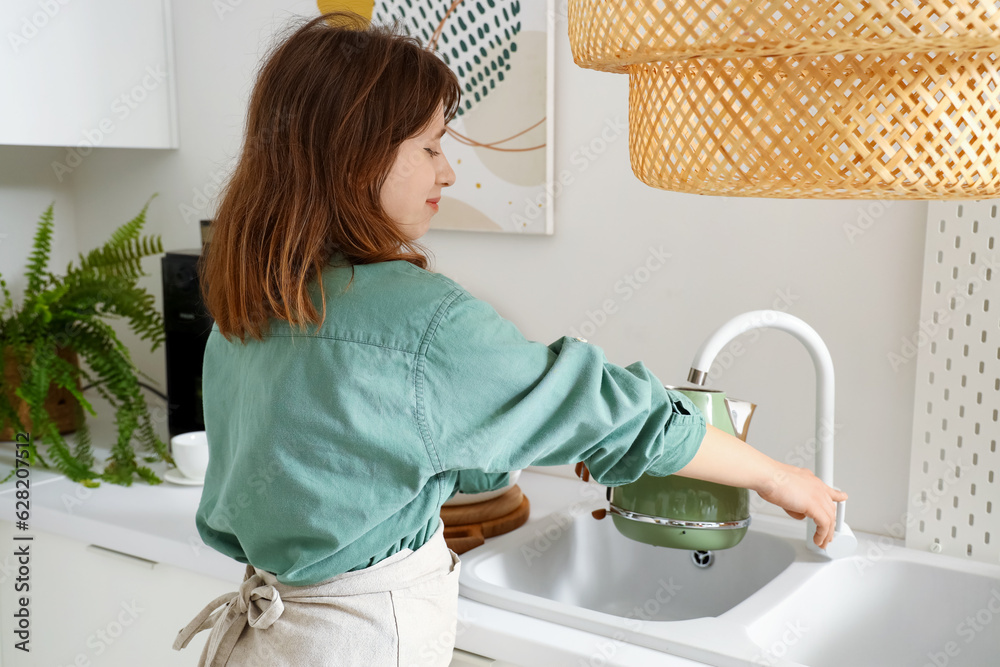 Young woman pouring water from faucet into electric kettle in modern kitchen