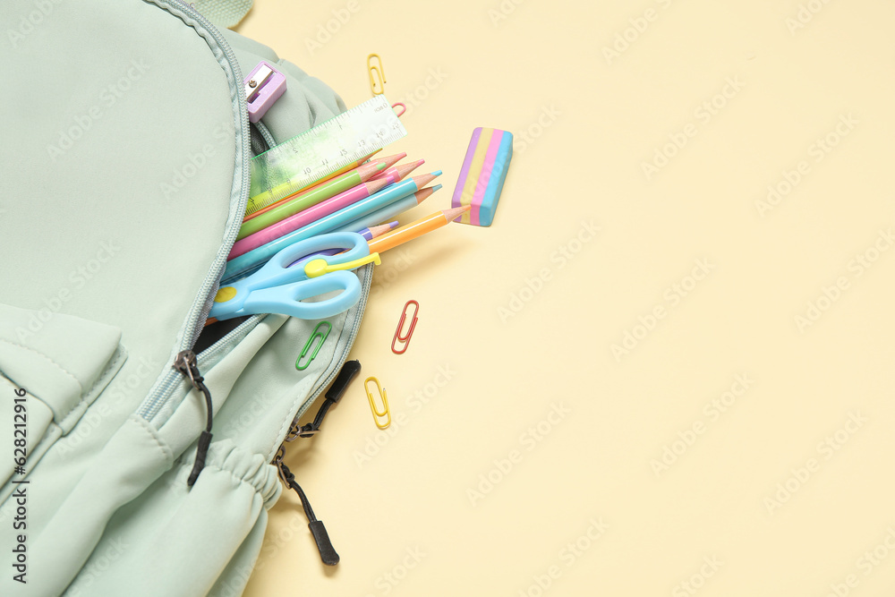 Green school backpack with different stationery on pale yellow background