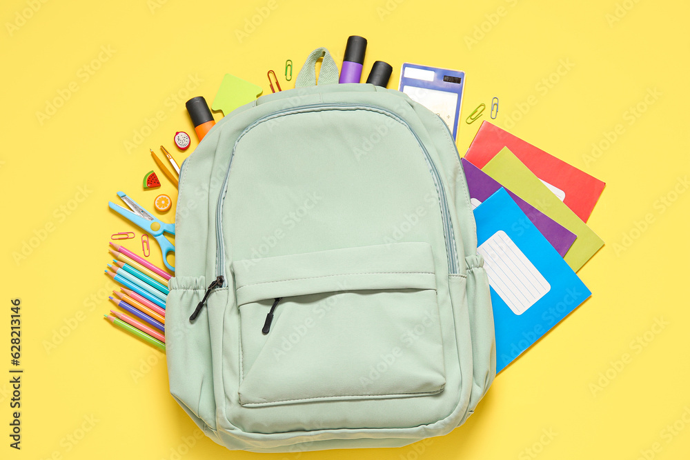 Green school backpack with different stationery on yellow background