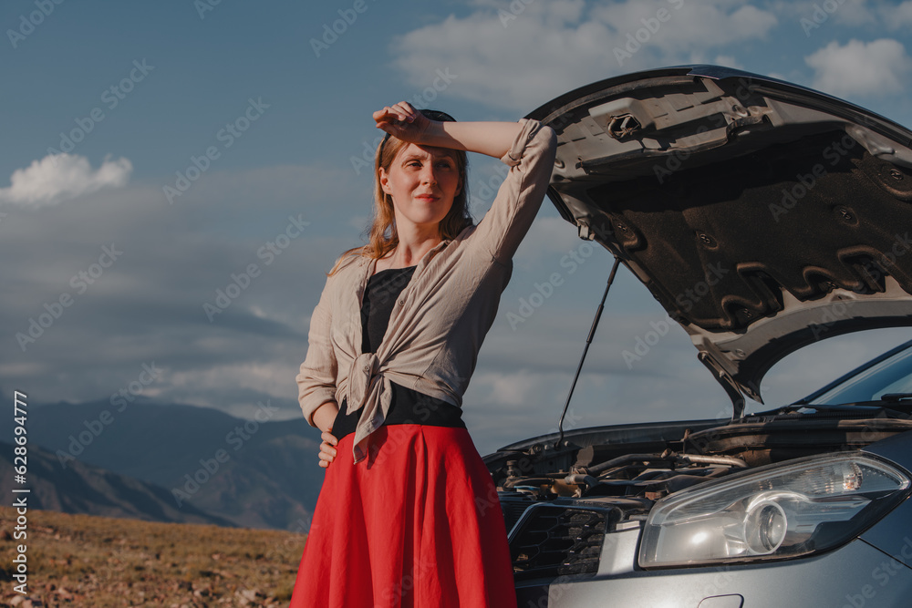 Tired young woman standing in front of broken down car on mountains background