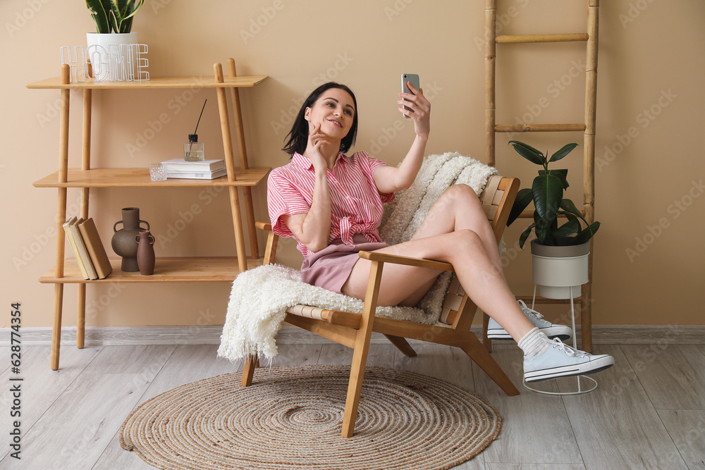 Beautiful woman with mobile phone taking selfie in wooden armchair at home