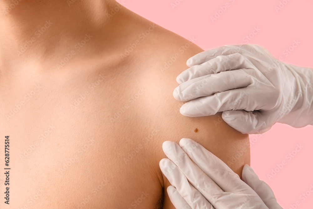 Dermatologist examining mole on young mans shoulder against pink background, closeup