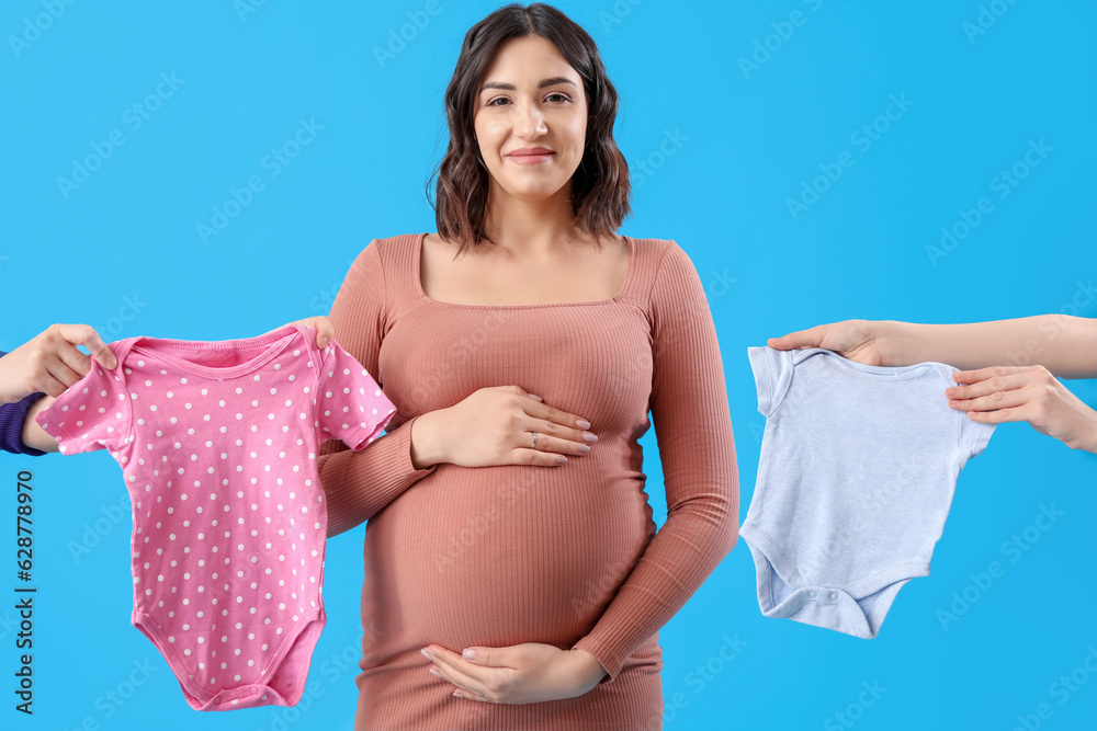 Young pregnant woman and female hands with baby bodysuits on blue background