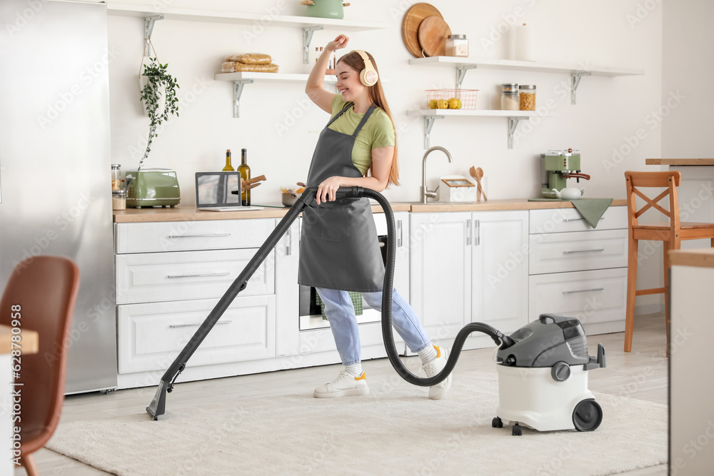 Young woman with headphones vacuuming carpet in kitchen