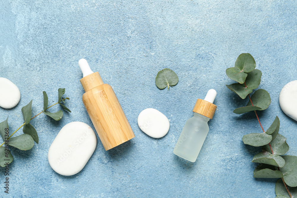 Bottles with cosmetic oil, eucalyptus branch and stones on blue textured background