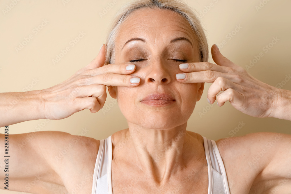 Sporty mature woman doing face building exercise on beige background, closeup