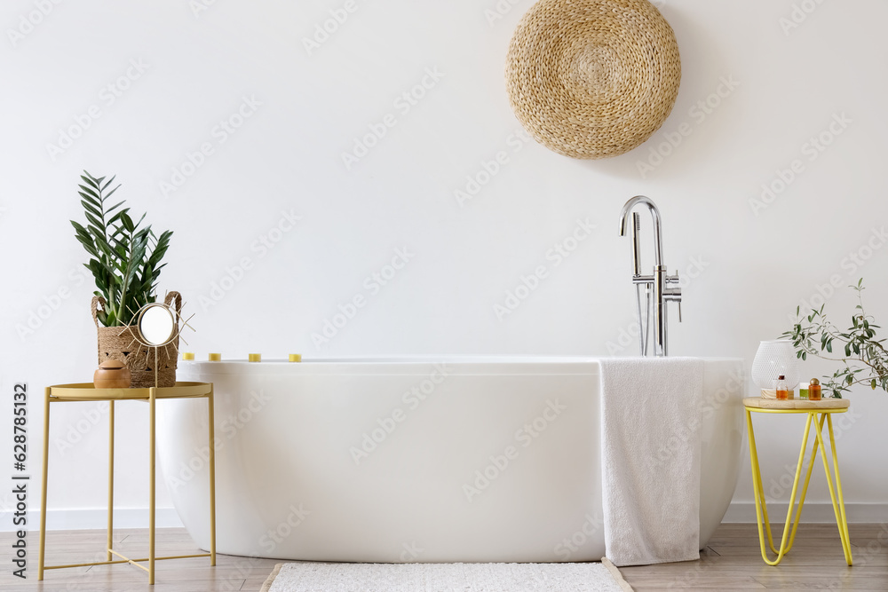 Interior of light bathroom with white bathtub, tables and houseplants