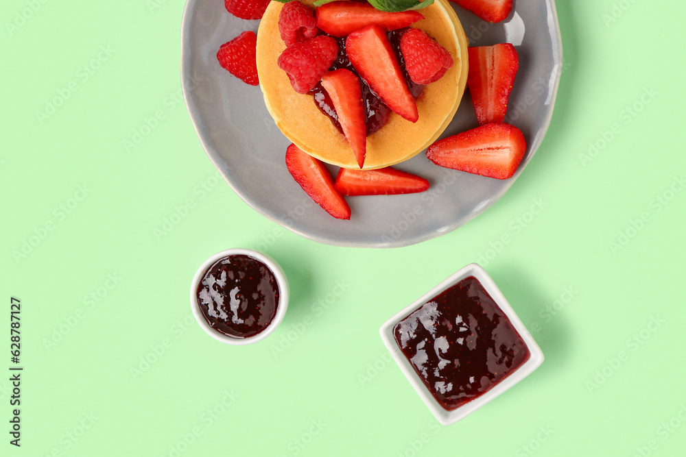 Plate with sweet pancakes, berries and jam on green background