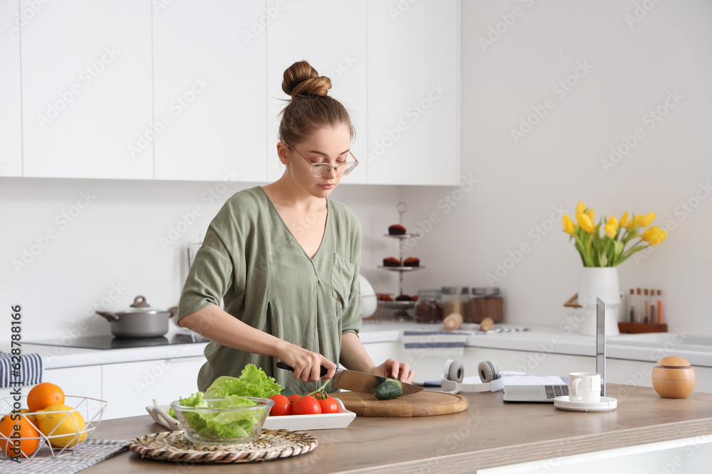 Beautiful young woman cutting cucumber at table with modern laptop in light kitchen