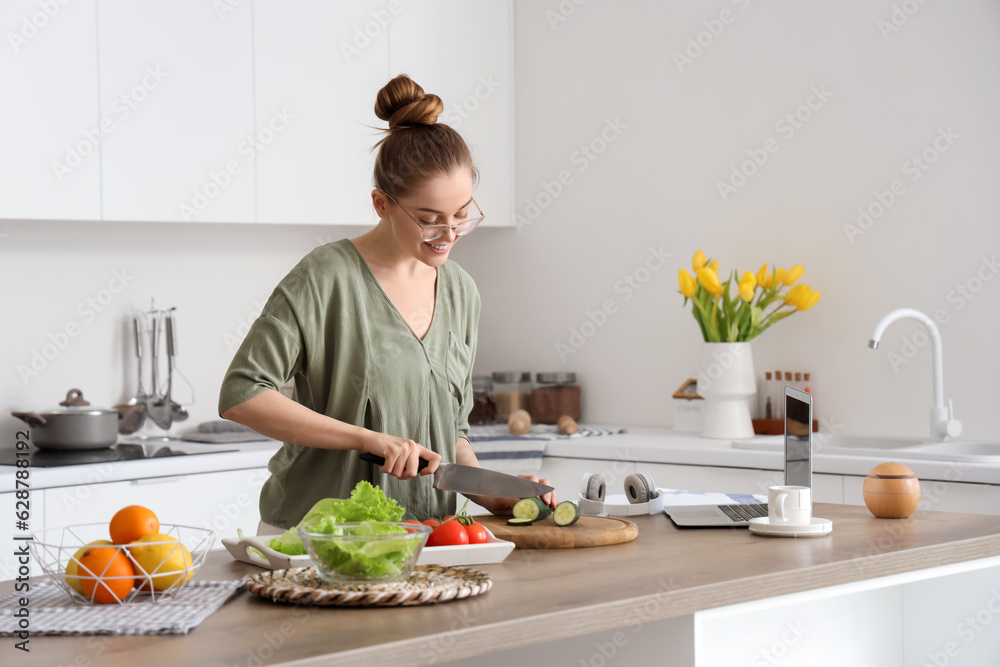 Beautiful young woman cutting cucumber at table with modern laptop in light kitchen