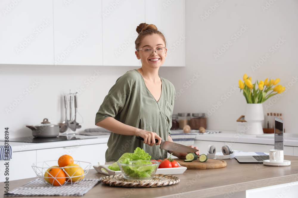 Beautiful young woman cutting cucumber at table with modern laptop in light kitchen