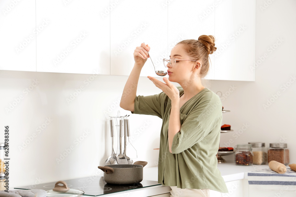 Pretty young woman tasting food from ladle in light kitchen