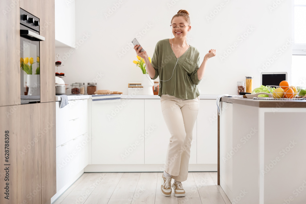 Happy young woman listening to music and dancing in light kitchen