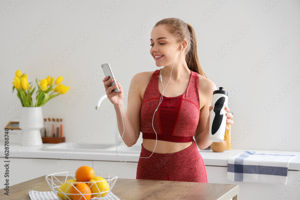 Sporty young woman with bottle of water listening to music in light kitchen