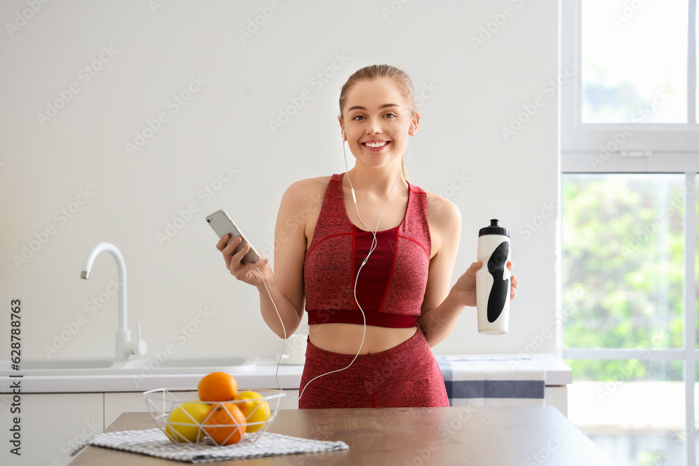 Sporty young woman with bottle of water listening to music in light kitchen