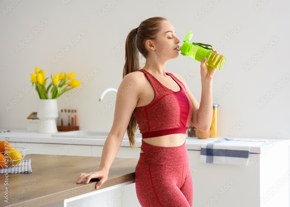 Sporty young woman drinking water from bottle in light kitchen