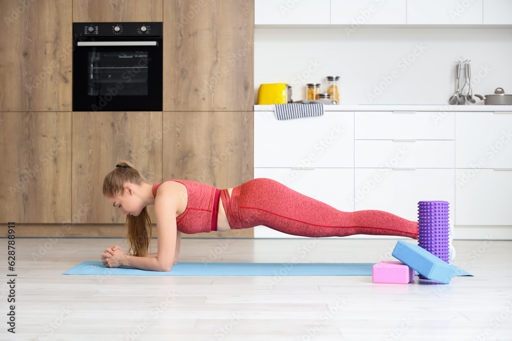 Sporty young woman exercising on yoga mat in light kitchen