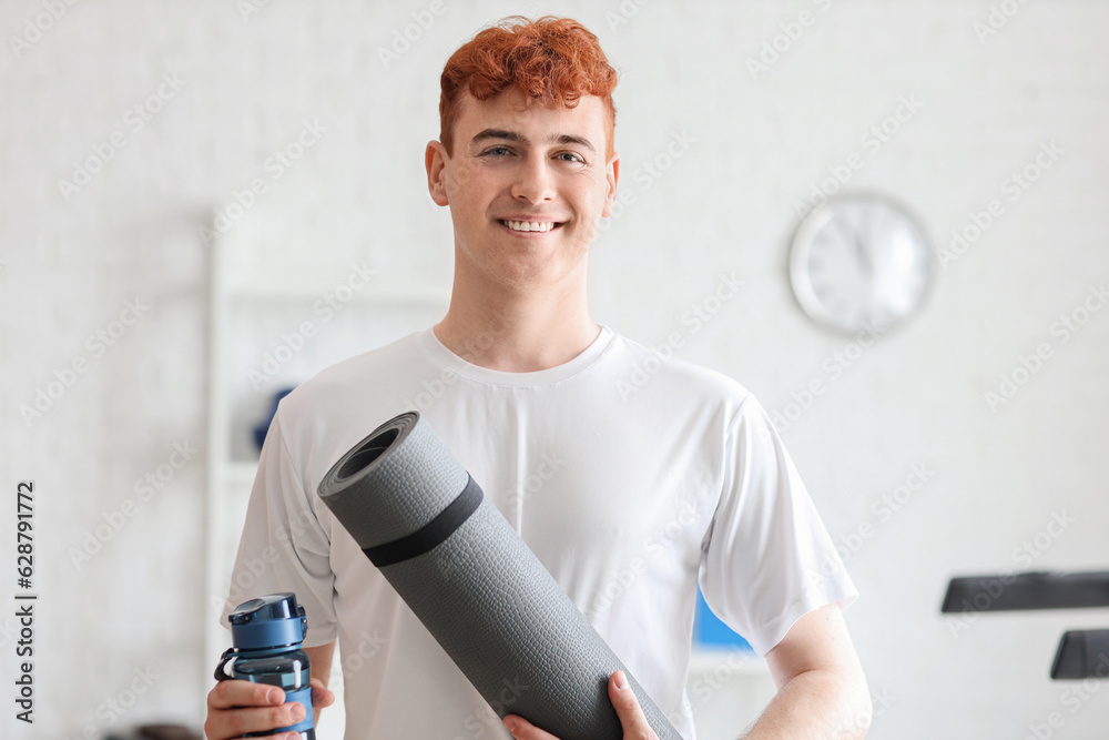 Young redhead man with sports water bottle and mat in gym