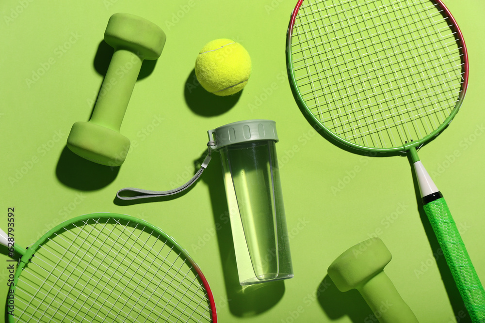 Composition with bottle of water, dumbbells, tennis rackets and ball on green background