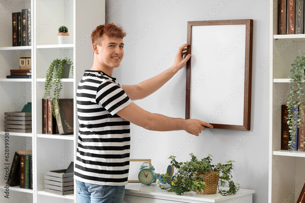 Young redhead man hanging blank frame on light wall at home