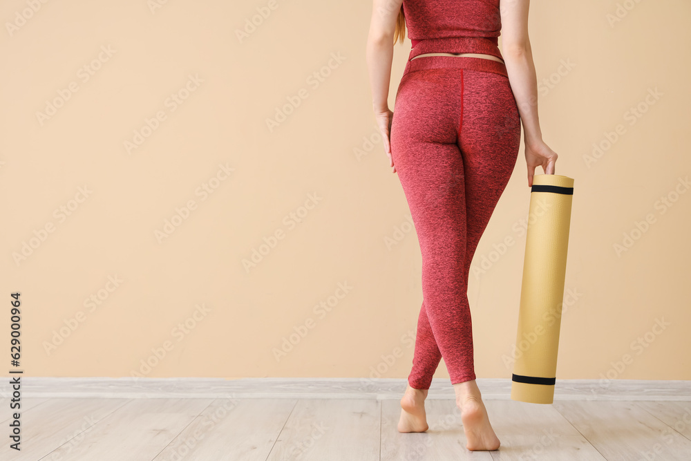 Sporty young woman in leggings and with yoga mat near beige wall, back view