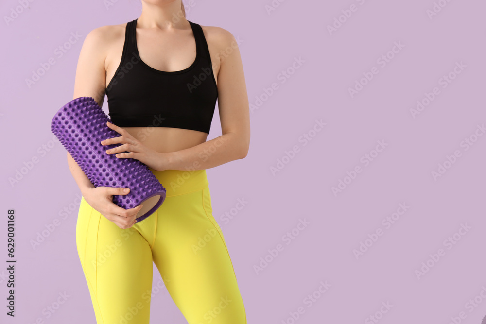 Young woman in sportswear and with foam roller on lilac background