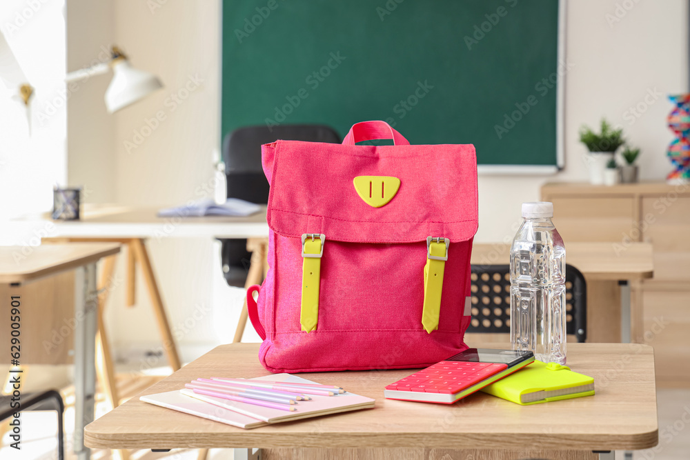 Pink school backpack with stationery and bottle of water on desk in classroom