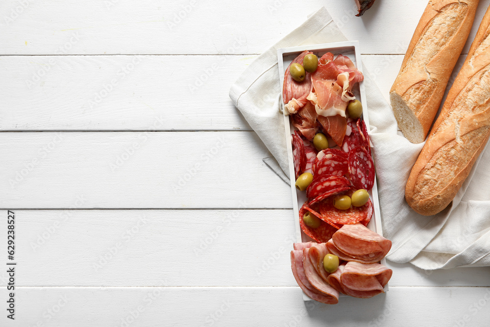 Plate with assortment of tasty deli meats and baguette on white wooden background